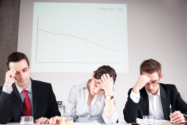 Three business professionals looking distressed at a table with a projected graph in the background showing a decline in sales over the year